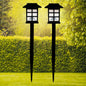 Two black solar garden lights with white paneled tops stand in a vibrant green lawn with hedges and trees.
