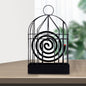 A black iron birdcage-style holder with a burning mosquito coil inside sits on a light wooden table.