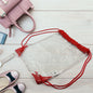 A clear ClassiCarry drawstring bag with red accents lies on a white wooden floor surrounded by a pink purse, phone, watch,