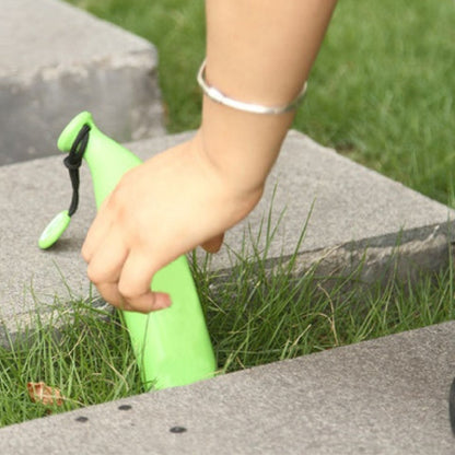 A hand reaches for a green banana-shaped umbrella case resting in green grass between concrete slabs.
