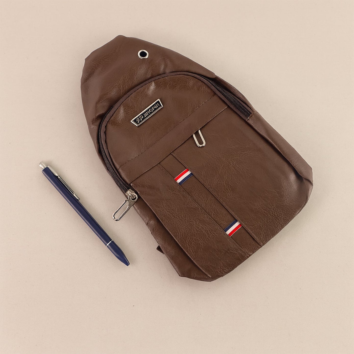 A brown leather sling bag lies flat on a light brown surface with a blue pen next to it.